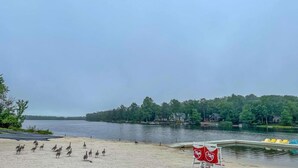 A lakeside view with a sandy beach, a dock with yellow chairs, and a red sign with a white symbol. - Spacious & Charming Poconos Pine Hideaway (Tobyhanna)