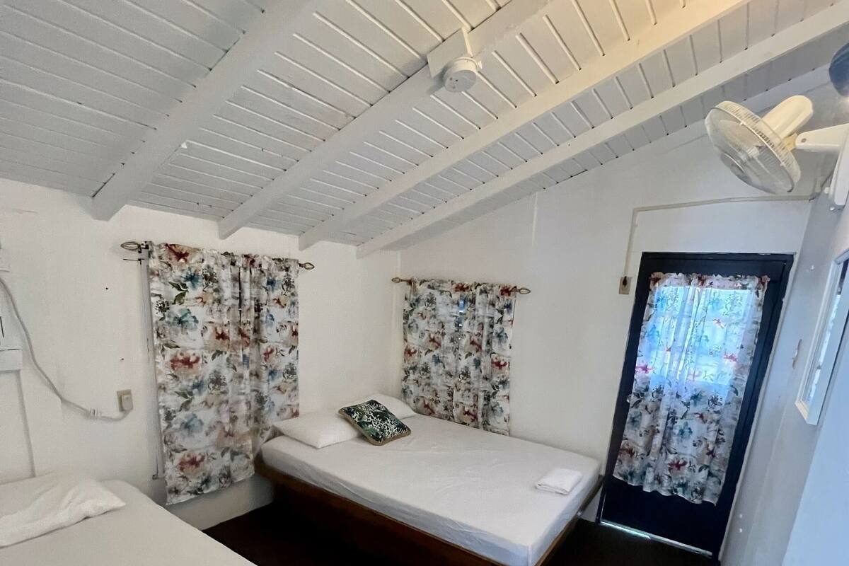 A small, compact room with two single beds, a ceiling fan, a door with a curtain, and a trash bin.