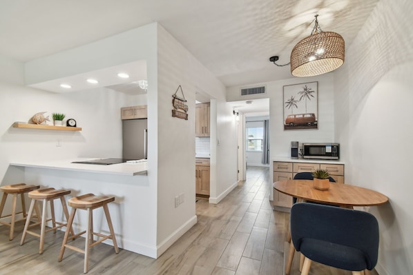 Una cocina moderna con una isla central, taburetes de madera, una mesa de comedor con sillas y una lámpara colgante.