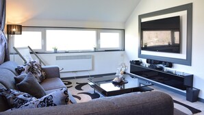 A modern living room with a sofa, coffee table, and a large flat-screen TV mounted on the wall. - Lake-view Apartment With Sauna, hot tub (Stavelot)