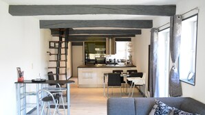 A modern living room with a grey sofa and patterned cushions, a dining area with a small table and chairs, and a kitchen in the background with stainless steel appliances and a wooden ceiling. - Lake-view Apartment With Sauna, hot tub (Stavelot)