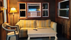 Een gezellige kamer met een houten plafond, een plafondventilator, een gestreept bankje, een tafeltje, een lamp en een raam met een televisie.