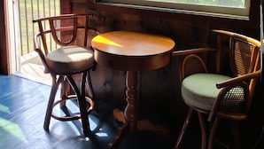 Een kamer met een houten interieur, een groot raam met uitzicht op bomen en een auto buiten, plus twee houten stoelen met kussens.