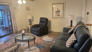 A living room with a black leather sofa, a glass coffee table, a black leather chair, a patterned rug, a ceiling fan, and a framed picture on the wall.