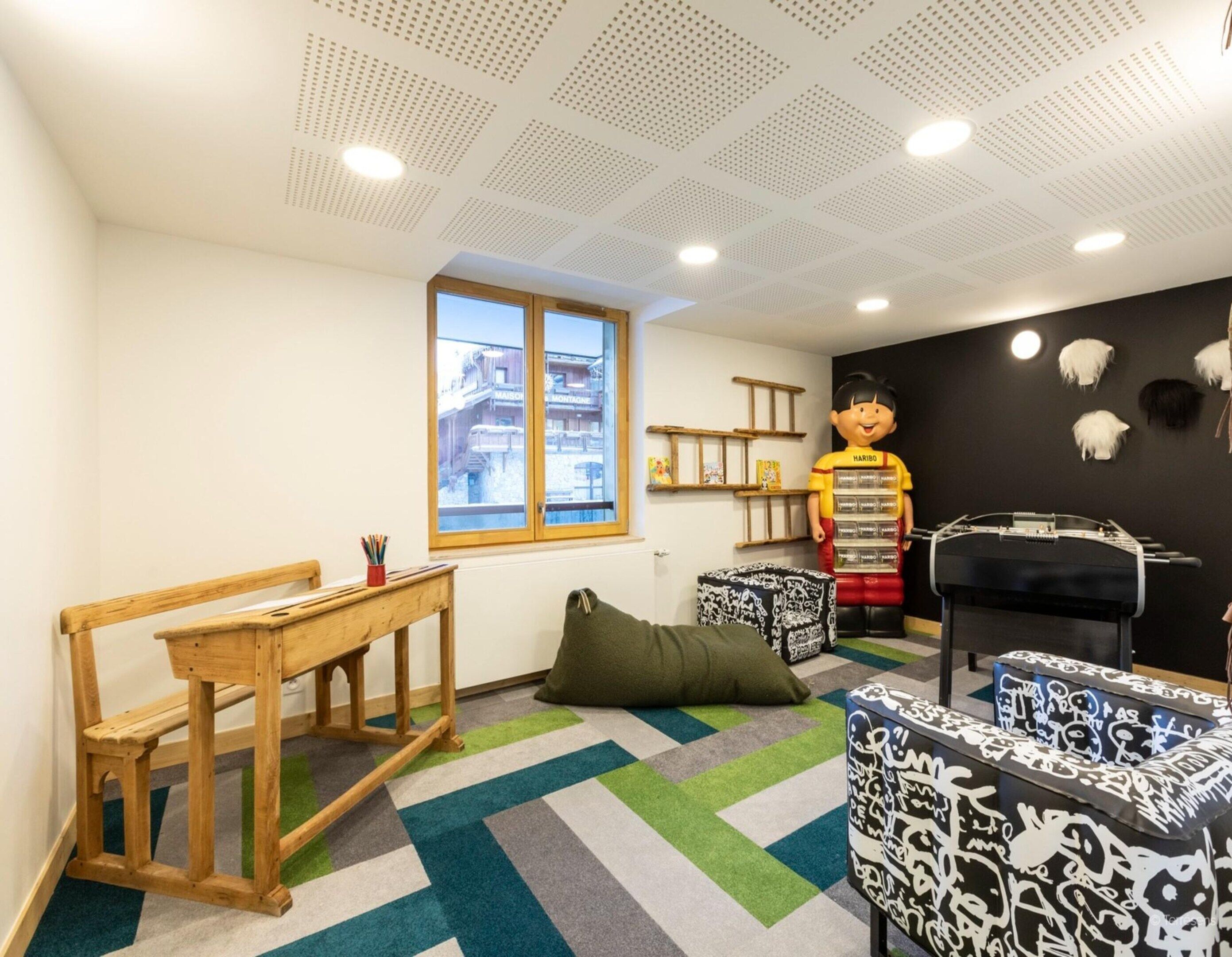 A children's playroom with a large window, a wooden bench, a green bean bag, and a black and white patterned ottoman.