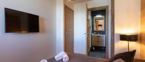 A bedroom with a bed, a TV mounted on the wall, a door leading to a bathroom, and a lamp.