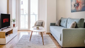 Ein modernes Wohnzimmer mit einem Flachbildfernseher, einem grauen Sofa, einem kleinen runden Tisch und einem großen Fenster mit Jalousien.