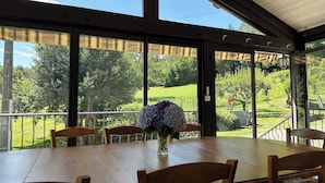 A covered patio with a wooden table and chairs, large windows offering a view of a green landscape.