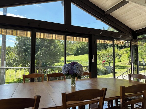 A covered patio with a wooden table and chairs, large windows offering a view of a green landscape.