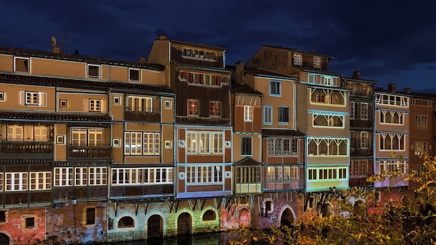 A nighttime view of a row of multi-story buildings with illuminated windows and balconies, reflecting on a body of water.