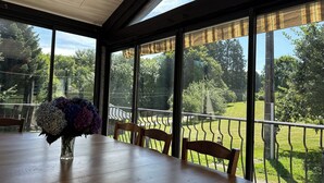 A dining area with a wooden table, chairs, and a vase of flowers. Large windows offer a view of a green landscape and a metal fence.