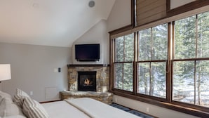 A bedroom with a stone fireplace, a large window with a forest view, and a flat-screen TV mounted above the fireplace. - 10 Trails Edge-Curate Telluride, Ski-in/out, Walk to Gondola, Market, Hot Tub (Mountain Village)