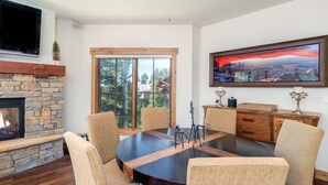 A dining area with a round table, chairs, a fireplace, and a large window with a view of trees and a house.