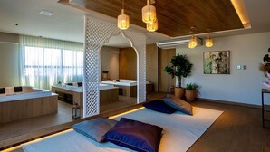 Een moderne hotelkamer met een houten plafond, een zithoek met kussens, een eettafel en een groot raam met dunne gordijnen.