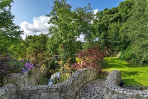 Un pont de pierre enjambe un bassin dans un jardin luxuriant, caractérisé par une végétation dense et un ciel dégagé.