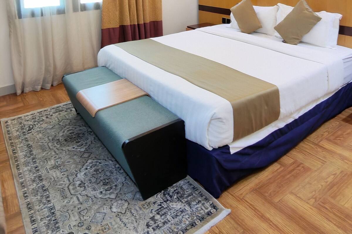 A hotel room with a large bed, a bedside table, a nightstand, a desk, a chair, a rug, and a window with curtains.