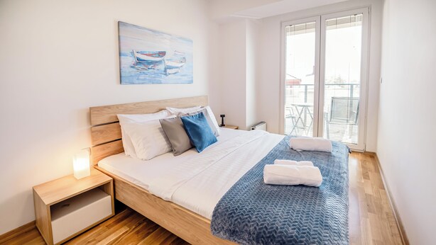 Cama bem arrumada com cabeceira de madeira, mesa de cabeceira com abajur e janela com persianas.