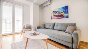 Uma sala de estar moderna com um sofá cinzento, uma mesa de centro branca e redonda e uma cadeira. Há um quadro de um barco na parede.