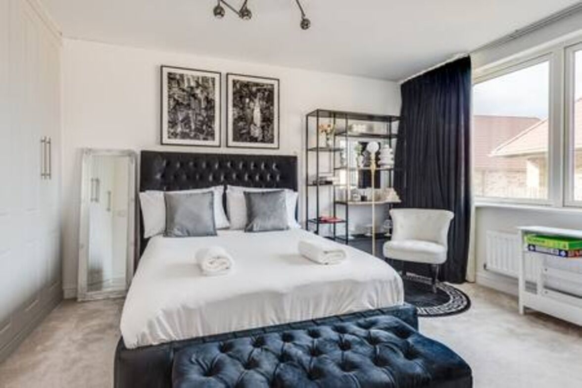 Une chambre à coucher moderne avec un grand lit, un pouf capitonné, une table de chevet et une fenêtre avec des rideaux.