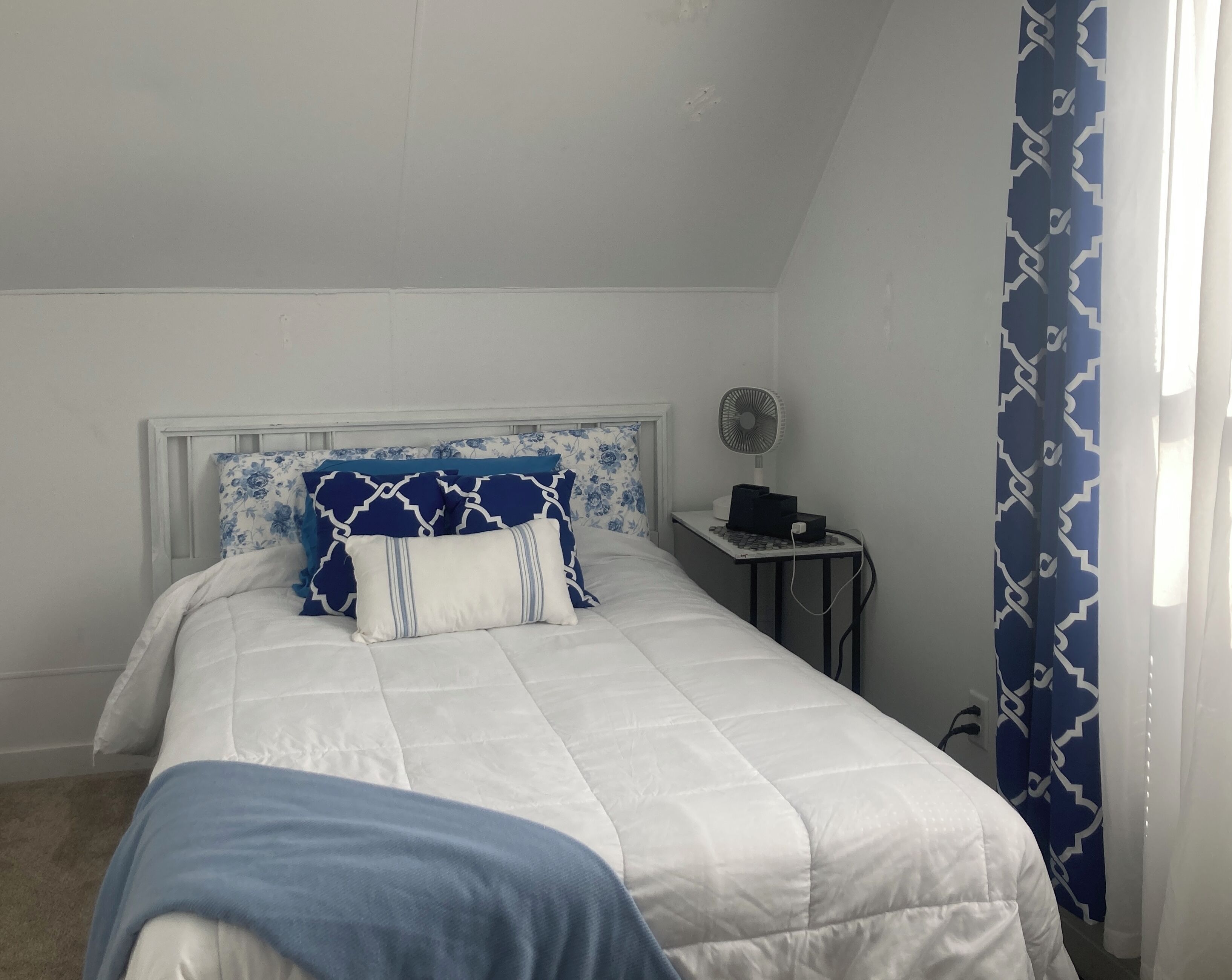 Un dormitorio con cama, mesita de noche y un ventilador, y una cortina estampada.