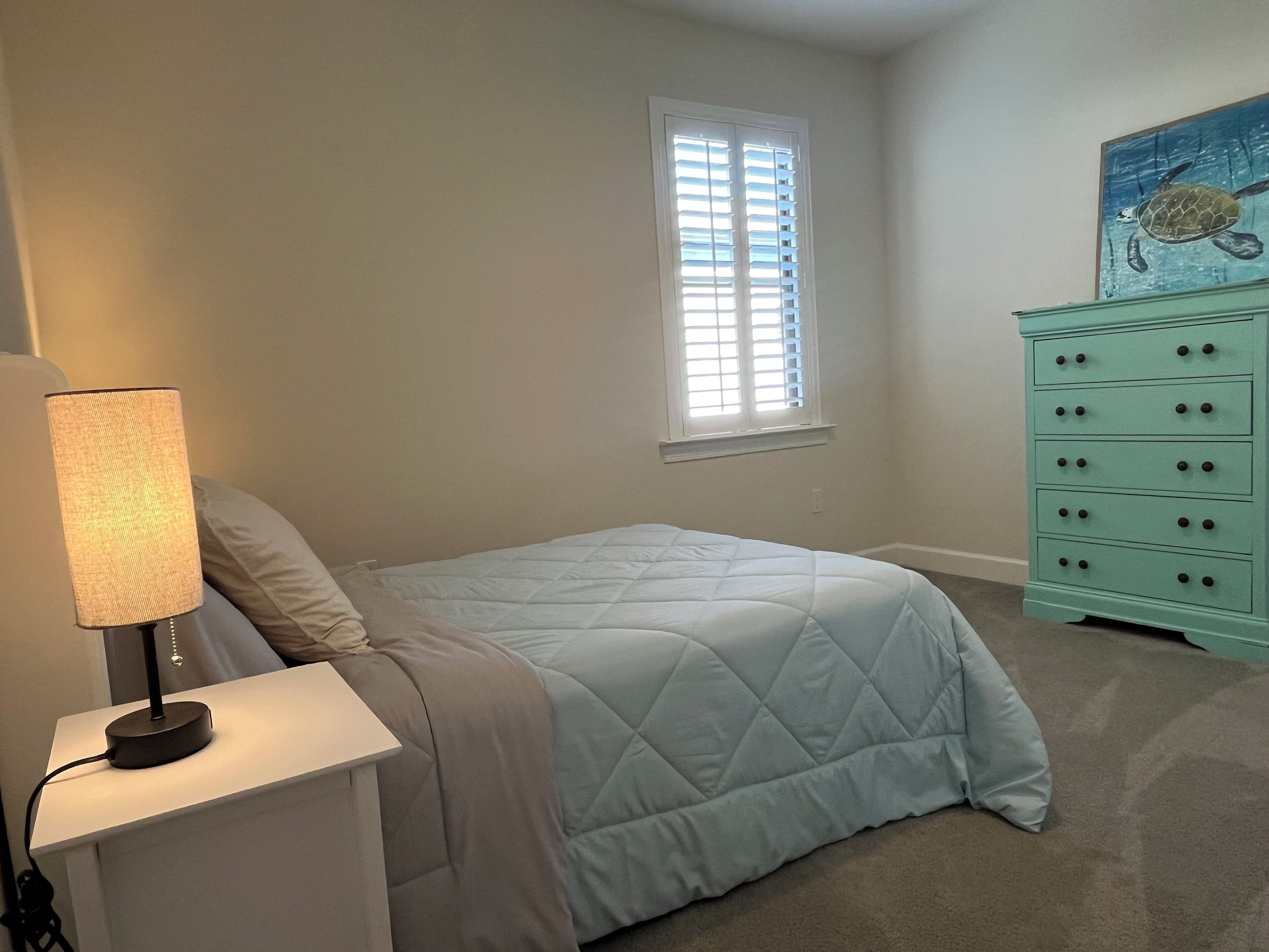 A bedroom with a bed, a nightstand, a lamp, a dresser, and a window with blinds.