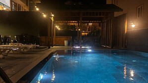 Een zwembad 's nachts, met een helder wateroppervlak, omgeven door een overdekt terras met zitplaatsen en een gebouw op de achtergrond.
