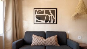 Um sofá cinzento com almofada estampada, parede bege e uma obra de arte abstrata emoldurada acima, com uma planta pendente à direita.