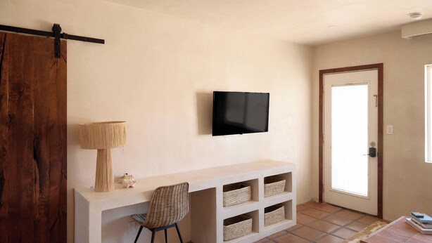 Quarto com cama, escrivaninha com cadeira, televisão e cestos organizadores.