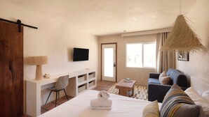 Quarto de hotel com cama, escrivaninha, poltrona, sofá, televisão e uma janela grande.