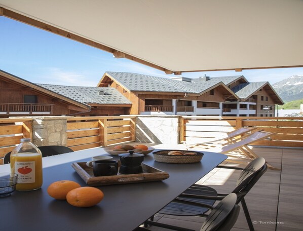 Una terraza de madera con una mesa dispuesta para el desayuno, incluyendo naranjas, una botella de zumo de naranja y una bandeja con tazas y un bol.