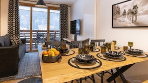 Un espace repas moderne avec une table en bois dressée pour quatre personnes, entourée de rideaux à motifs noirs et blancs, d’un canapé noir et d’un grand tableau encadré représentant des loups accroché au mur.