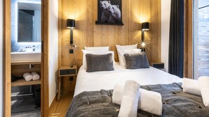 Une chambre d’hôtel moderne avec une tête de lit en bois, un grand lit avec une literie blanche et grise, deux lampes de chevet, un tableau encadré représentant des ours polaires et une vue sur un paysage enneigé par la fenêtre.