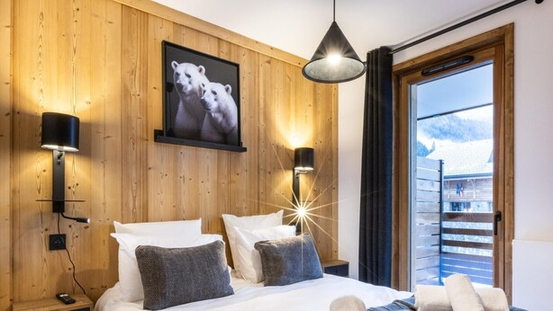 Une chambre d’hôtel aux murs lambrissés, un lit recouvert de draps blancs, deux lampes fixées au mur, un tableau représentant des ours polaires et une vue sur une montagne, visible à travers une porte coulissante.