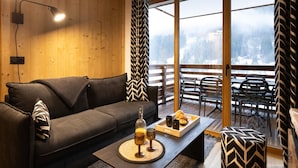 Un salon moderne avec un canapé, une table basse sur laquelle sont disposés des boissons et des pâtisseries, une grande fenêtre donnant sur un paysage enneigé et un rideau à motifs.