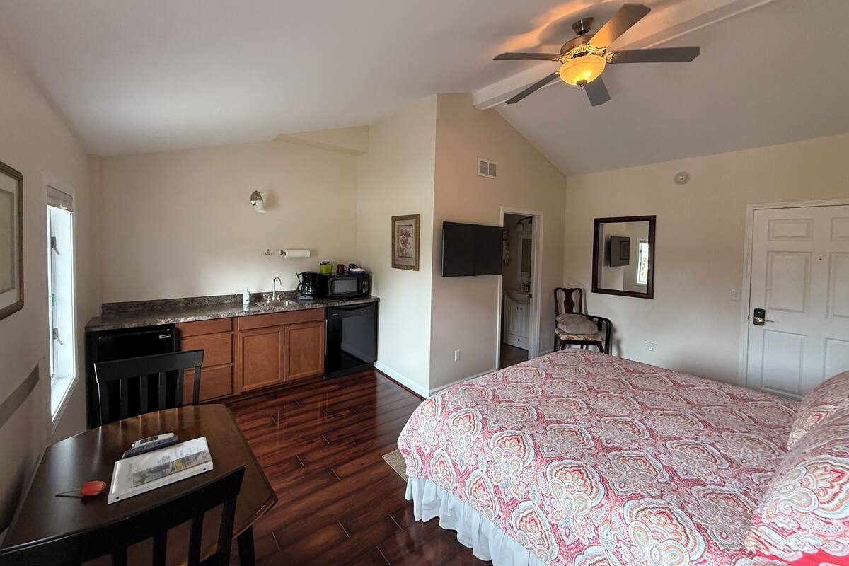 A bedroom with a bed, a desk, a chair, a ceiling fan, a TV, and a kitchenette with a microwave and sink.