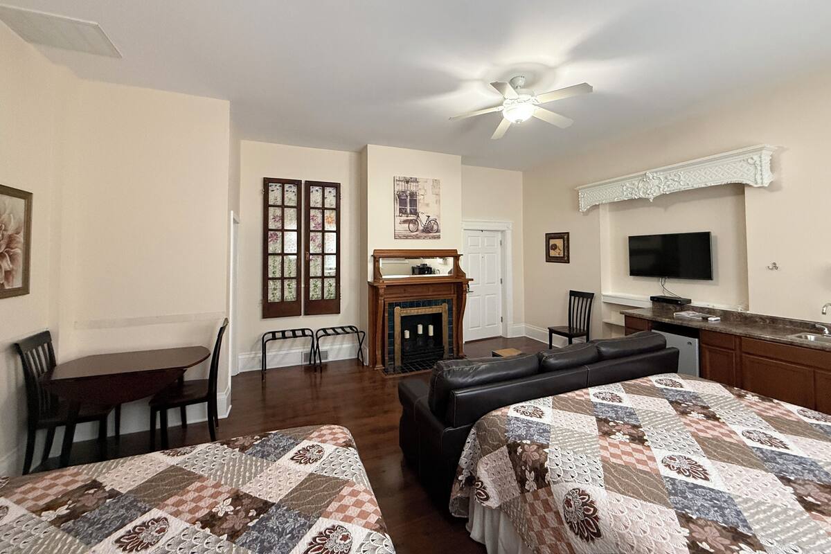 A room with two beds, a dining table, chairs, a fireplace, a television, and a kitchen area.