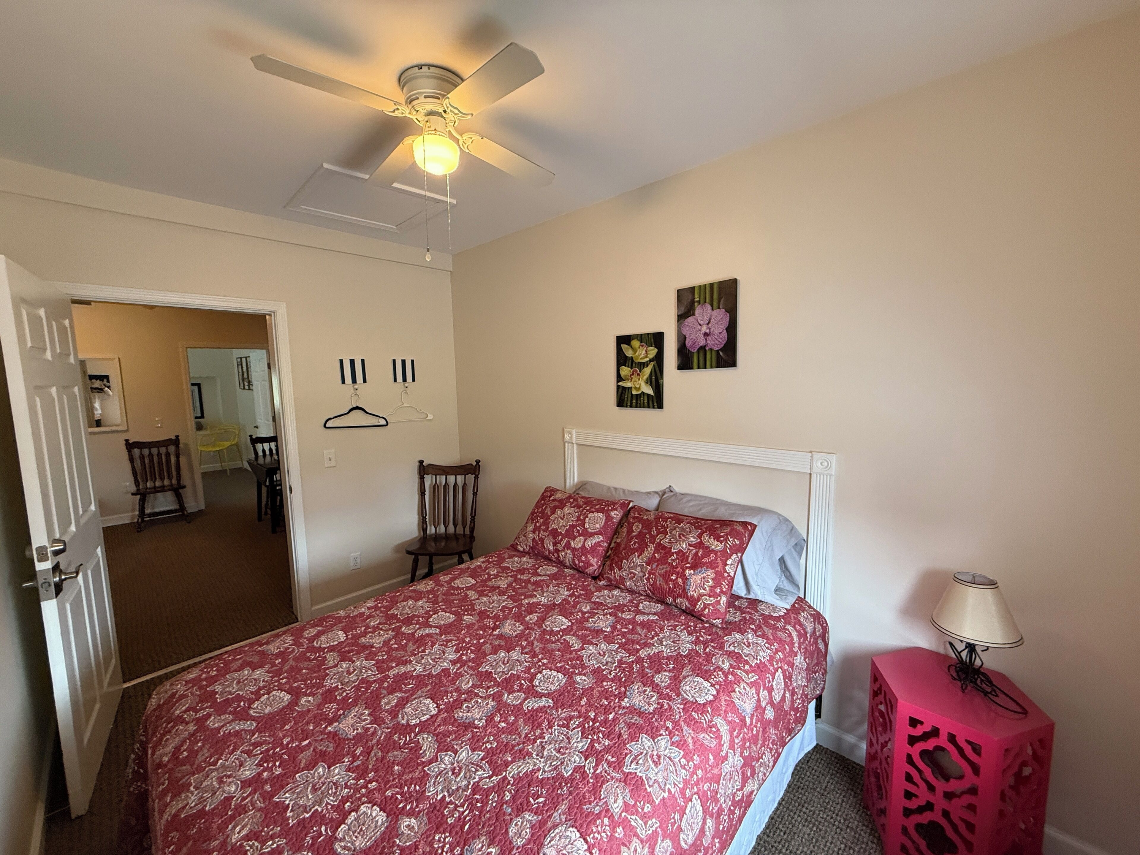 A bedroom with a bed, a ceiling fan, a nightstand, and a door leading to another room.