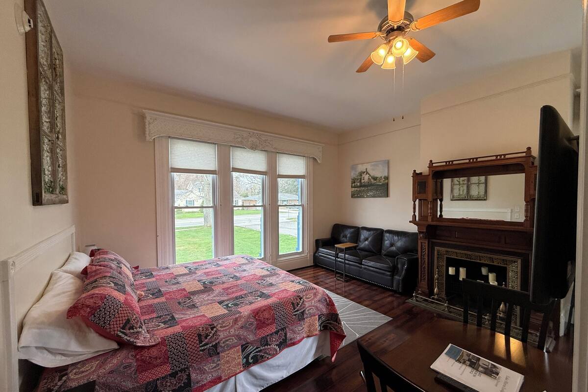 A bedroom with a bed, a desk, a chair, a sofa, a fireplace, and a ceiling fan.