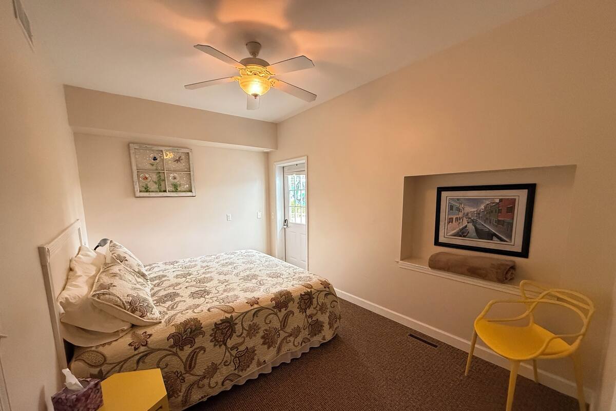 A bedroom with a bed, a ceiling fan, a window, a door, a framed picture, a yellow chair, and a small table with a tissue box and remote control.