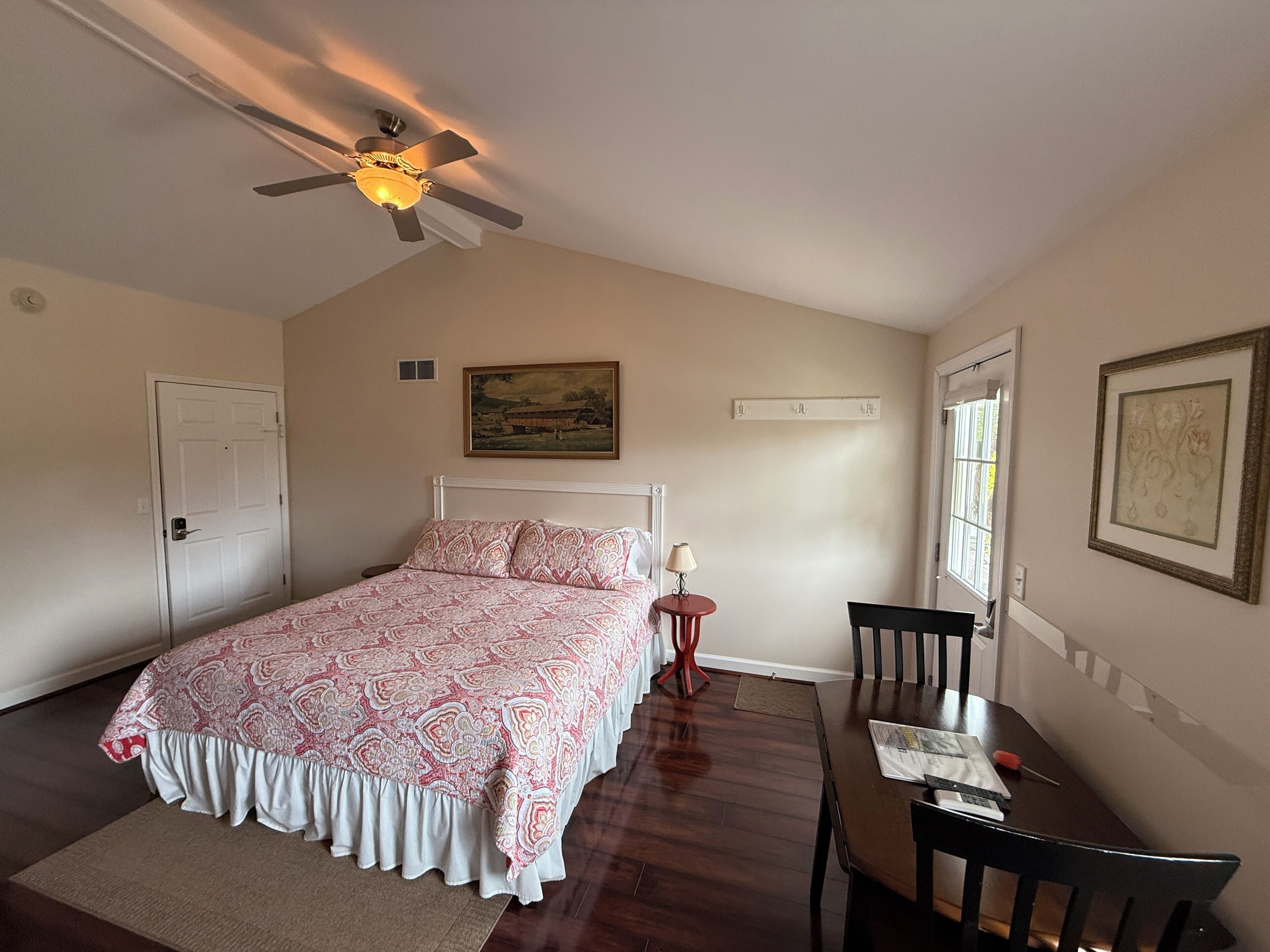Une chambre à coucher comprenant un lit, un ventilateur de plafond, un bureau avec une chaise, une fenêtre et une porte.