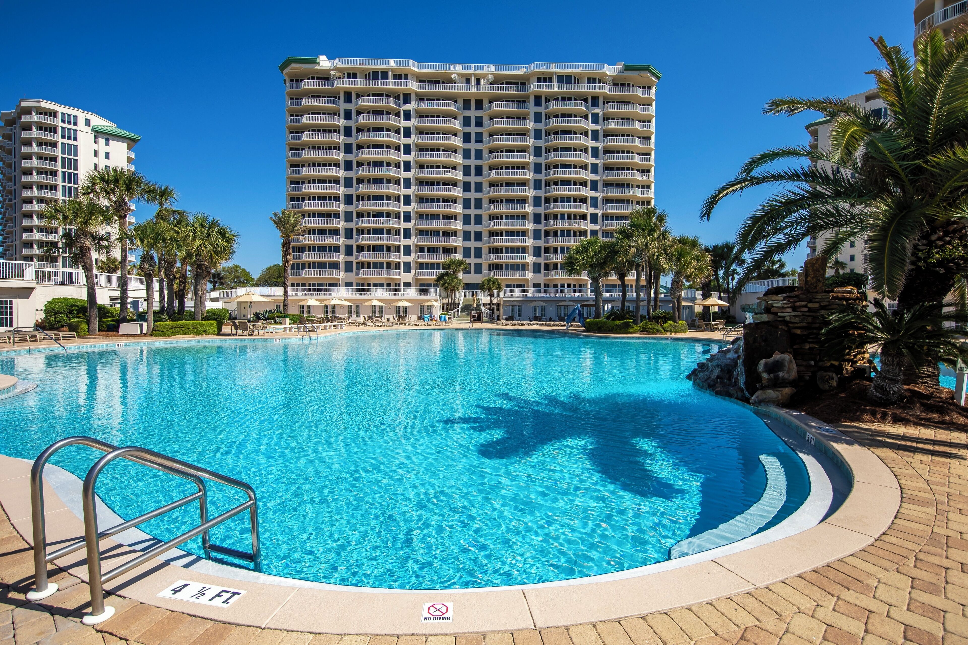 A swimming pool with a metal ladder, surrounded by palm trees and a large hotel building in the background.