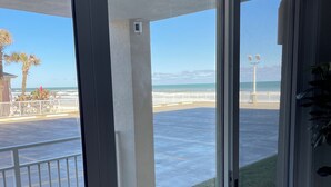 Une vue depuis un balcon avec une porte vitrée, donnant sur une plage bordée d’une clôture blanche, des palmiers et un ciel dégagé.