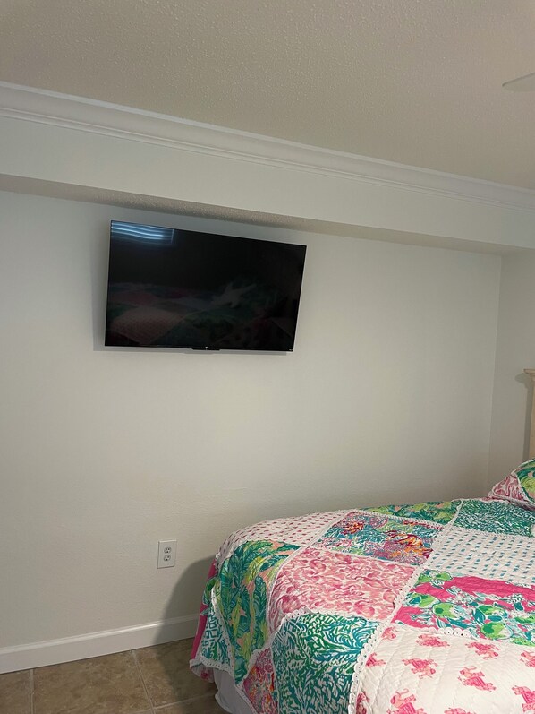 A bedroom with a bed covered in a colorful quilt, a television mounted on the wall, and a white ceiling.
