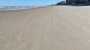 Une vaste plage de sable où l’on peut voir des empreintes de pas qui mènent vers l’océan. Des immeubles de grande hauteur se dressent sur le côté droit de la plage.