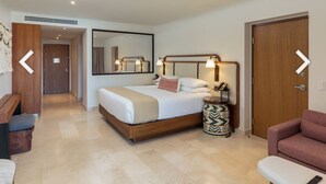 A modern hotel room with a large bed, bedside table, and a mirror. There is a door leading to another room and a bench in the corner.