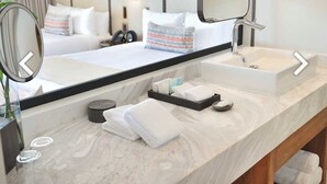 A hotel bathroom with a large mirror, two sinks, a vanity, and a bed with white linens.