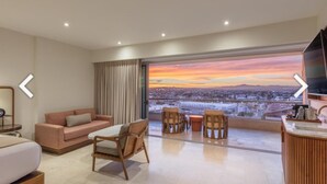A modern hotel room with a large window offering a city view, a sofa, two armchairs, a small table, and a TV.