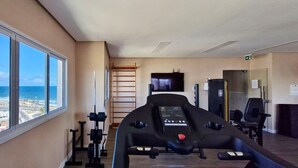 Un gimnasio con cinta de correr, banco de pesas y mancuernas. Una ventana ofrece vistas al océano y al paisaje urbano.