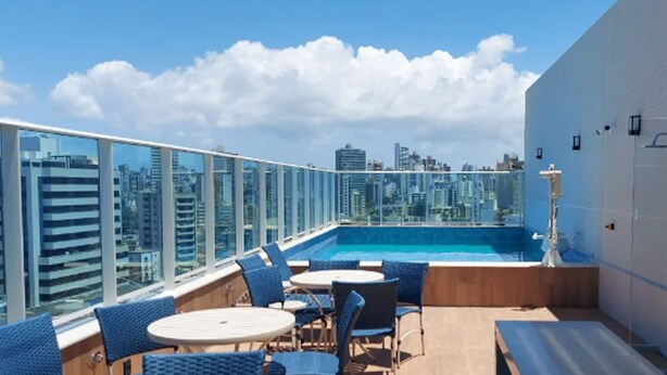 Un área de piscina en la azotea con barandas de vidrio, sillas azules y vistas a la ciudad.