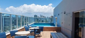 Ein Poolbereich auf dem Dach mit Glasgeländern, blauen Liegestühlen und Blick auf die Stadt.